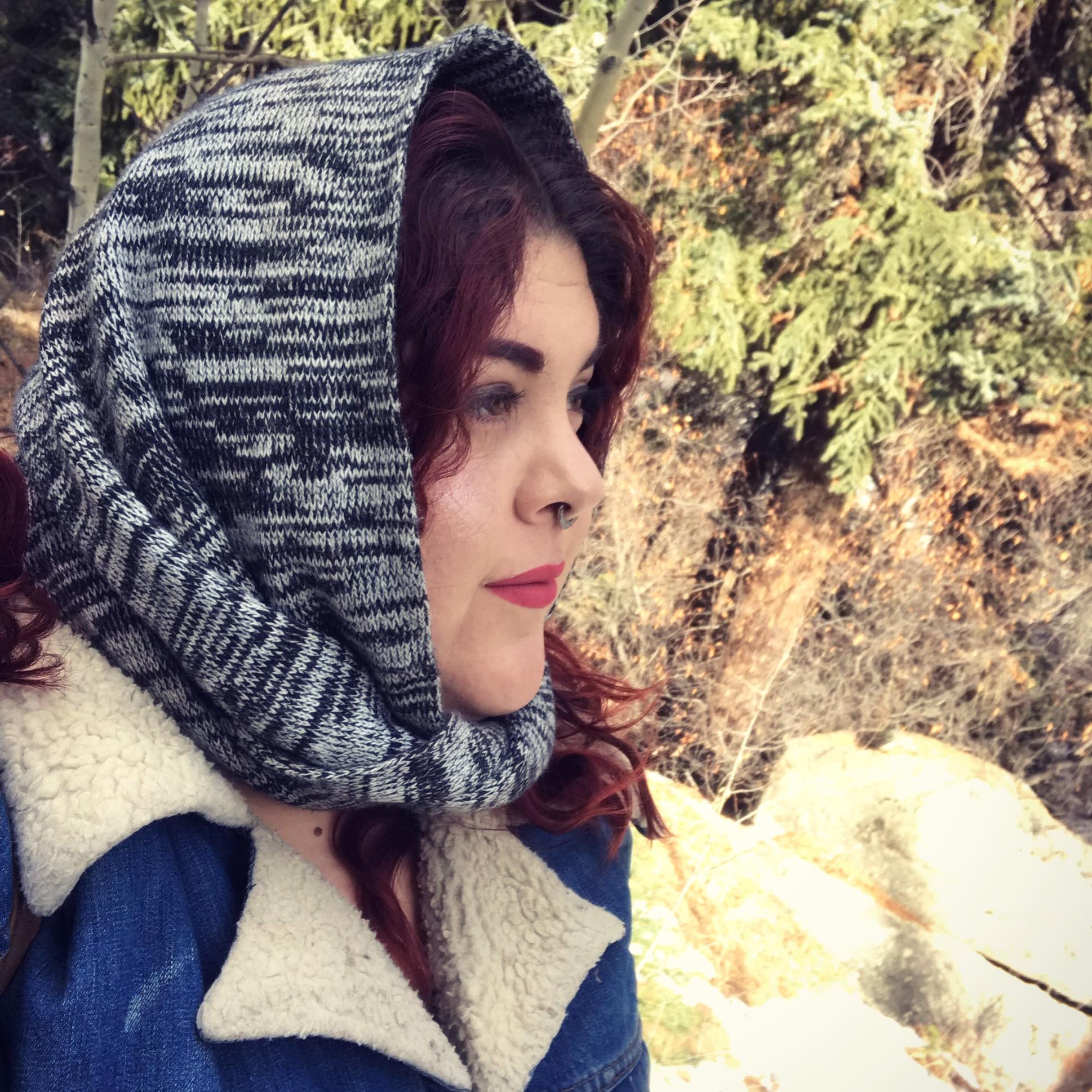 Author Victoria Parra looks to the right, red hair covered in a black and grey scarf. She is standing in front of trees.